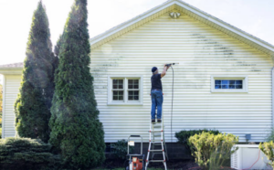 Older man on a ladder, power washing the side of a house.