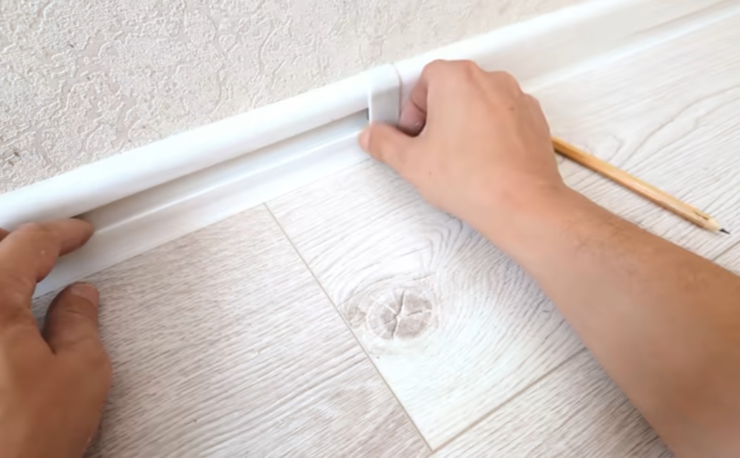 A handyman fitting white plastic baseboard molding along a room wall.