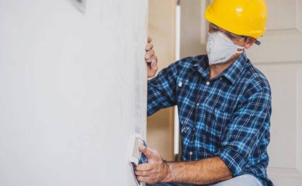 Drywall Repair Services: Smoothing Wall Surface with Safety Gear in Residential Home Worker wearing safety gear smoothing a wall surface during Drywall Repair Services inside a residential home.