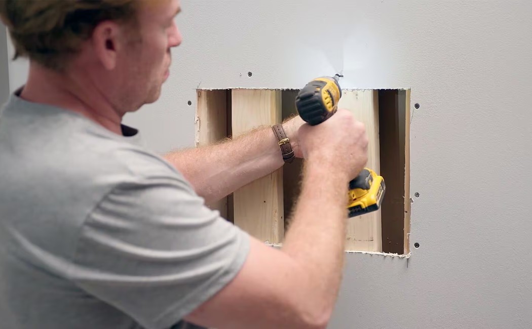 Man using a drill to fix wooden studs inside a wall opening, illustrating Drywall Repair Services.