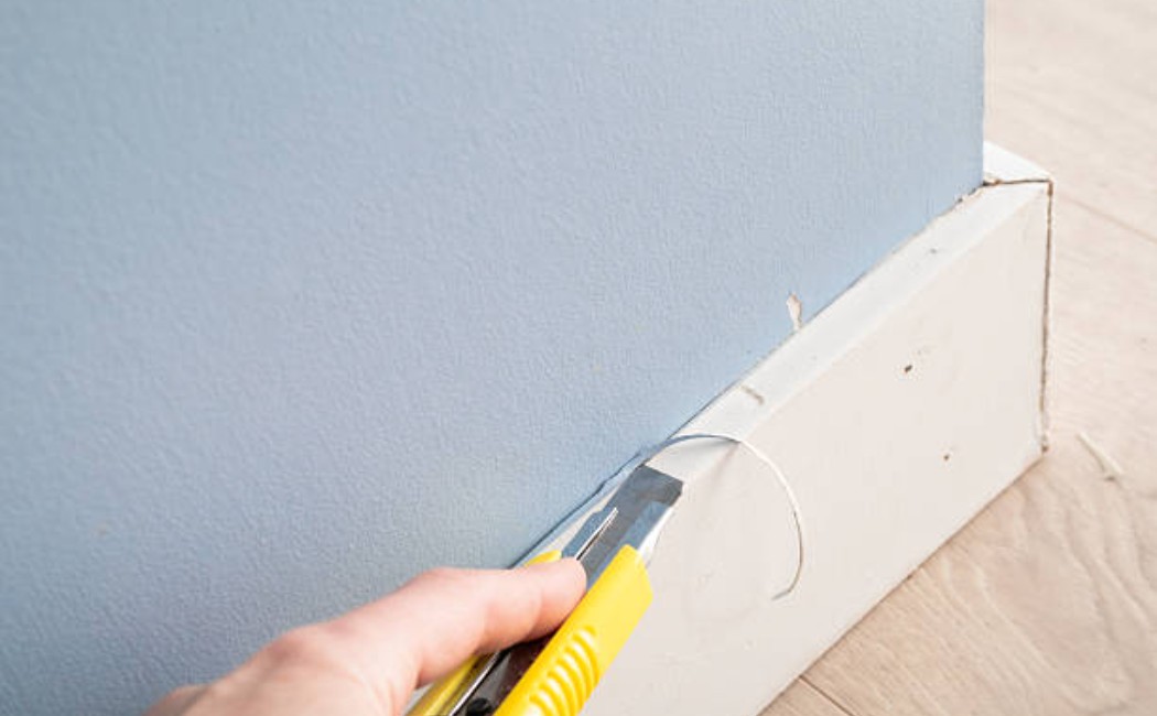 Hand using a utility knife to trim and repair wall trim along the floor, illustrating professional baseboard services.