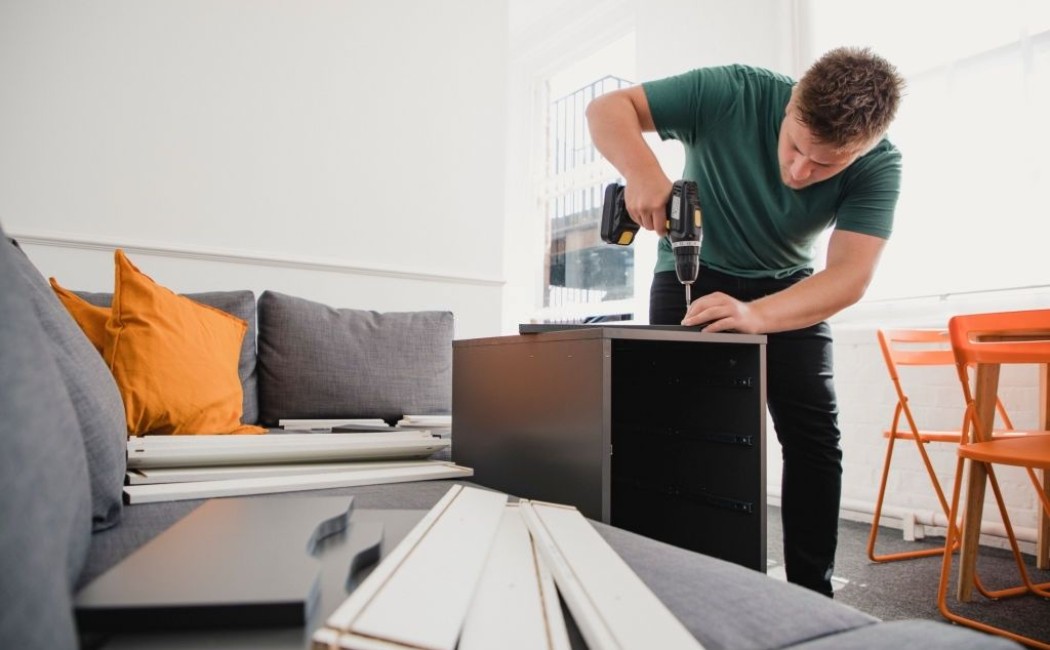 Man Assembling Black Cabinet Using Power Drill DIY. A man uses a power drill to assemble a black cabinet in a bright living room with furniture parts spread around him.